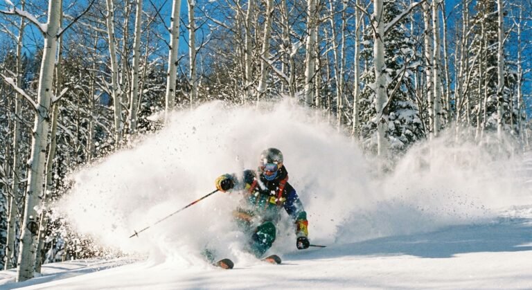 Nuage de poudreuse sèche (Champagne Powder) soulevé par un skieur à Aspen, Colorado, avec une forêt de trembles (Aspen trees) blancs en arrière-plan.