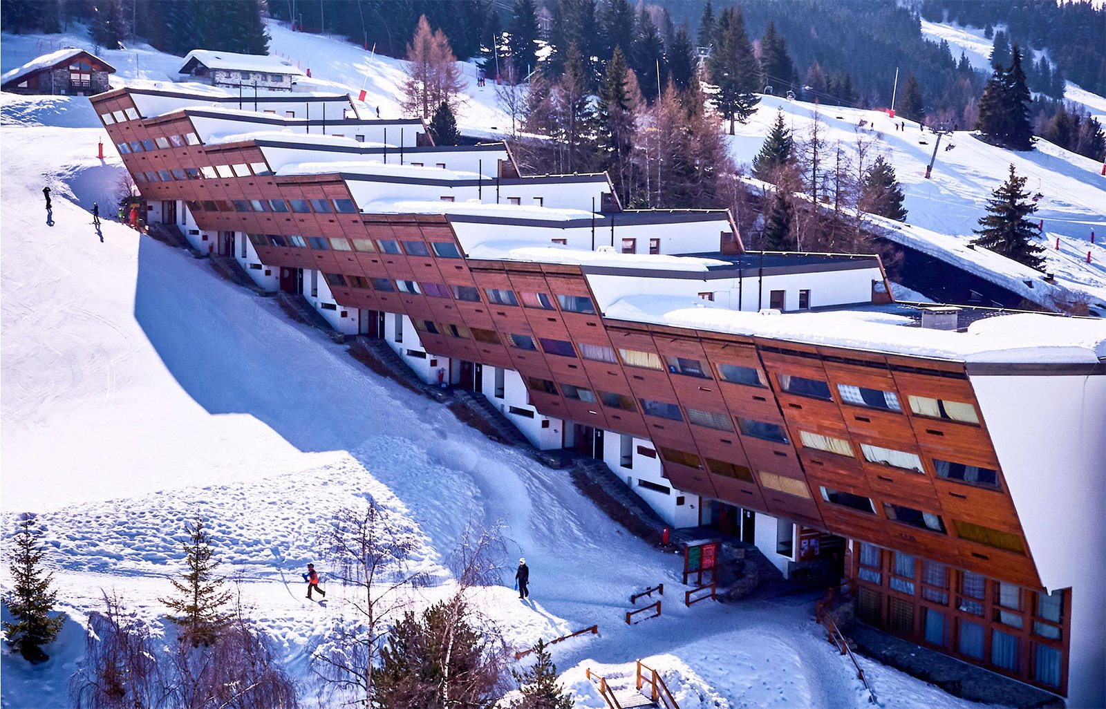 Bâtiment aux lignes modernes et aux toits plats recouvert de neige, épousant parfaitement la pente naturelle de la montagne dans les Alpes.