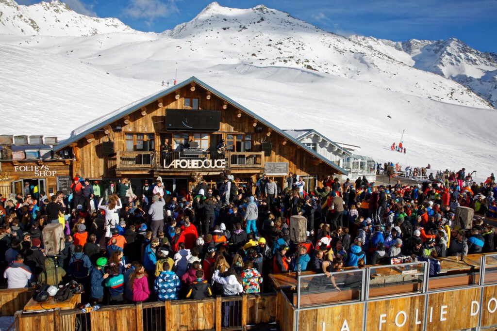Foule de skieurs célébrant la fin de journée en musique avec des boissons, illustrant l'ambiance festive des clubs à ciel ouvert sur les pistes.
