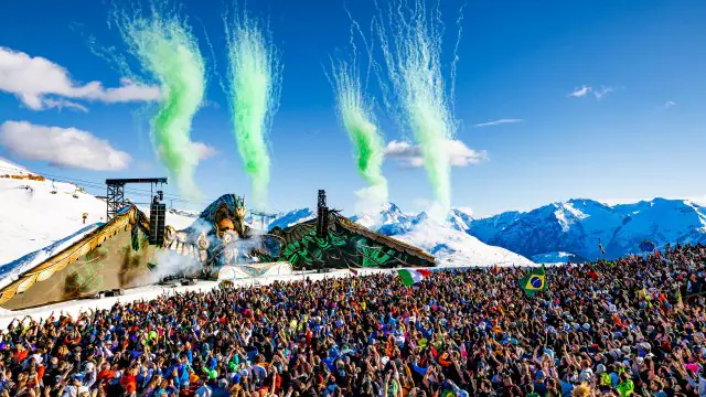 Foule de festivaliers dansant sur la neige sous un grand soleil printanier, vêtus de tenues colorées et de lunettes de soleil.