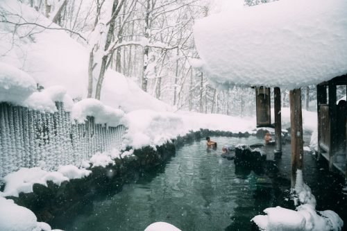 Un onsen traditionnel (bain thermal japonais) en extérieur, l'eau brûlante contrastant avec les rochers sombres et la neige fraîche.