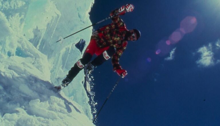 Cliché d'archive sépia de Patrick Vallençant en pleine action, engagé dans une pente vertigineuse avec ses longs skis droits et un piolet à la main.