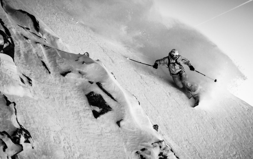 Un télémarkeur moderne, genou plié au ras du sol, fendant un nuage de neige poudreuse profonde avec une esthétique parfaite.