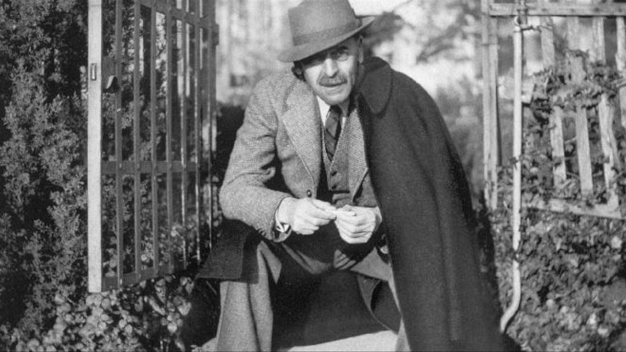 Homme moustachu en chapeau accroupi devant une grille de jardin